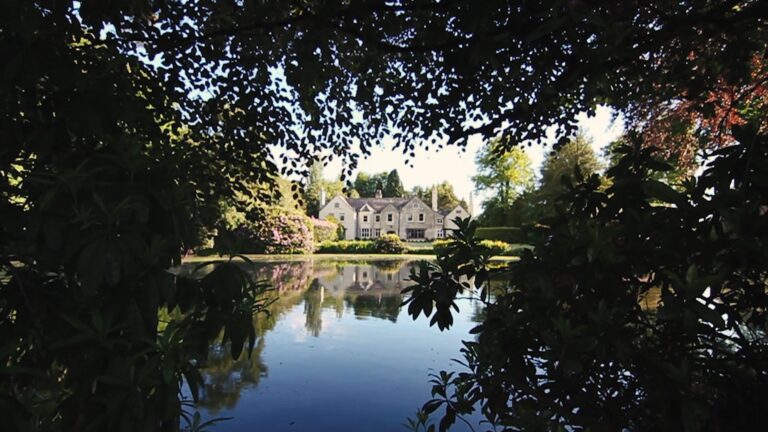 Moor Hall’s main house reflected in the lake, framed by garden foliage.